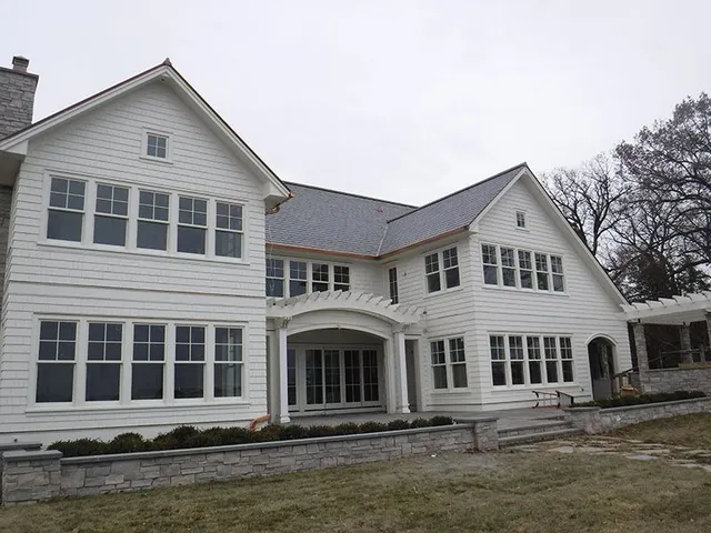 White two-story house with many windows and a stone wall at the base. Overcast day.