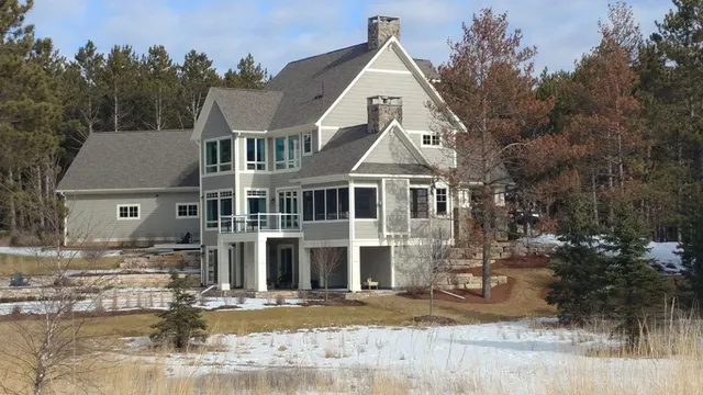 Two-story gray house with multiple windows and balconies, set in a snowy, wooded area.