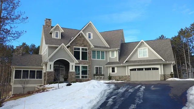 A large gray house with stone accents, a snow-covered driveway, and a blue sky.