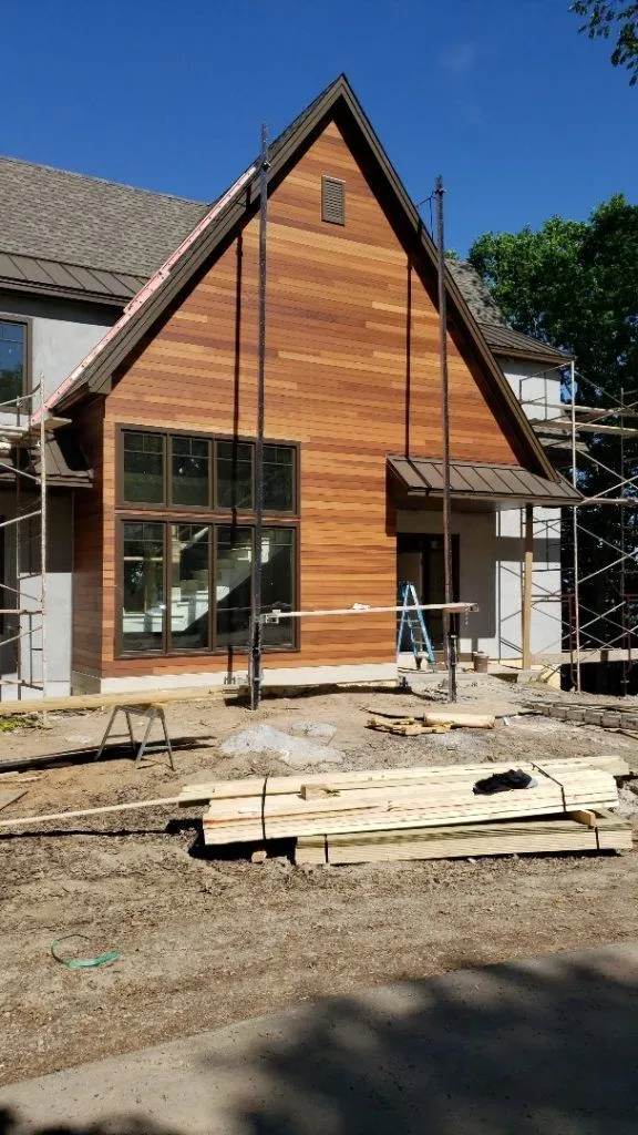 House under construction; exterior wood siding, large windows, scaffolding, and lumber in foreground.